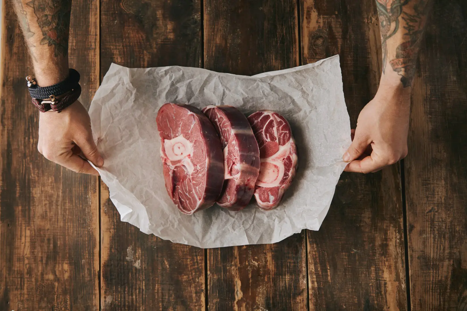 top view tattooed hands hold white craft paper with three raw great leg steaks with bone it everything old wooden brushed table